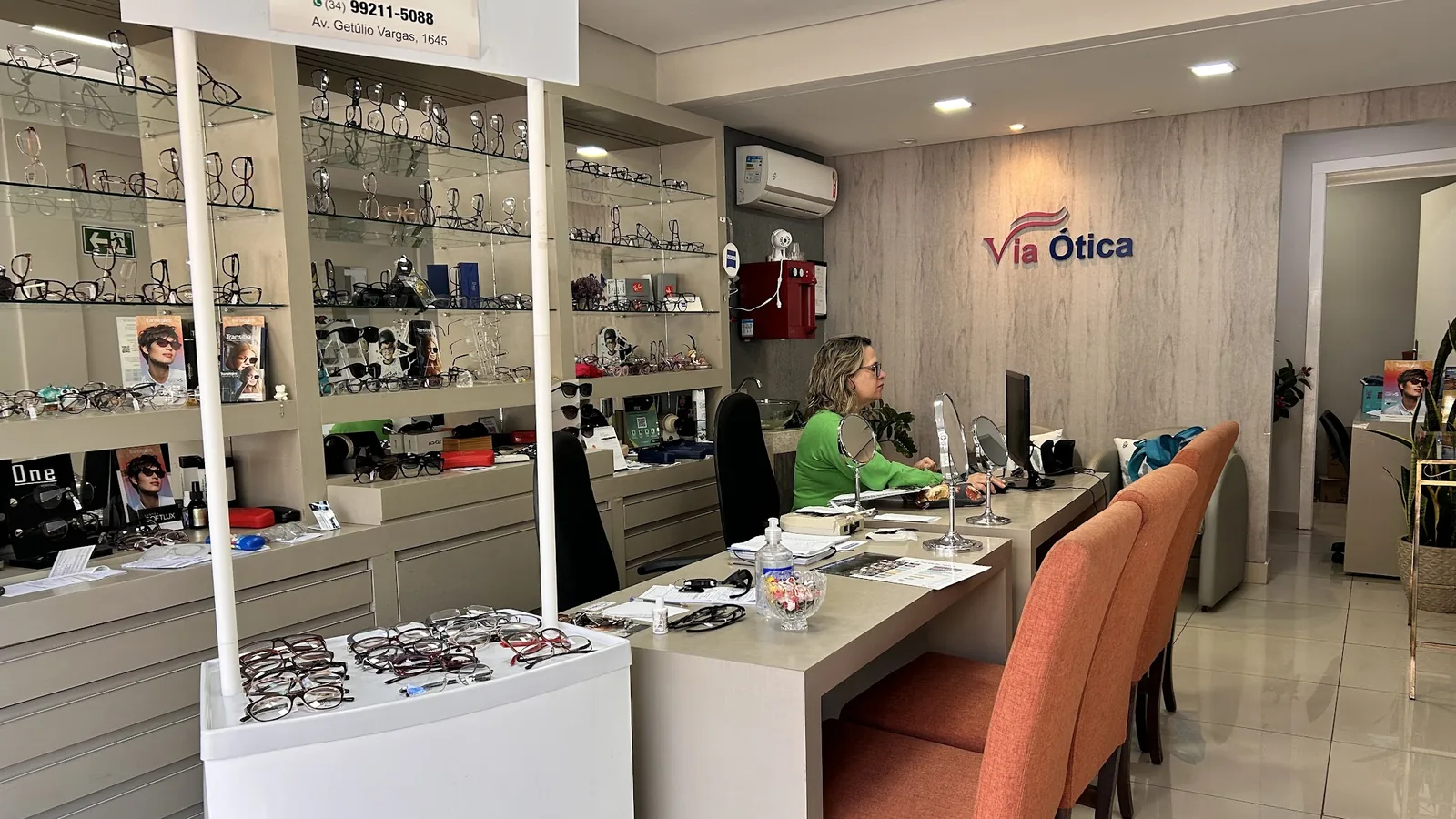 Interior da Via Ótica: parede de armações de grau e solar, balcão de atendimento e a placa com o endereço.